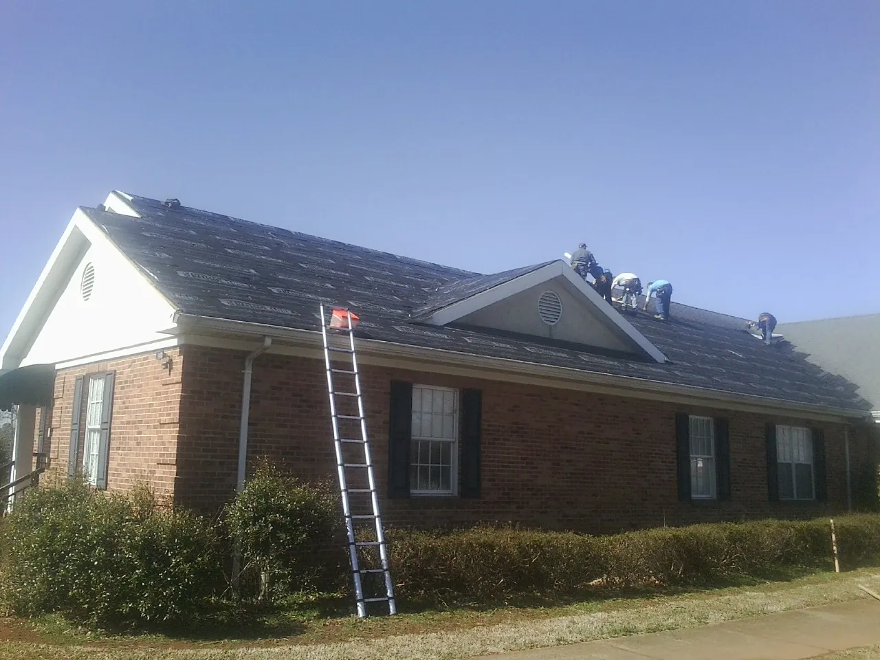 Crew tearing shingles off a brick ranch roof with a ladder set — Metal Roof Repair work by our crew in Thetford Center