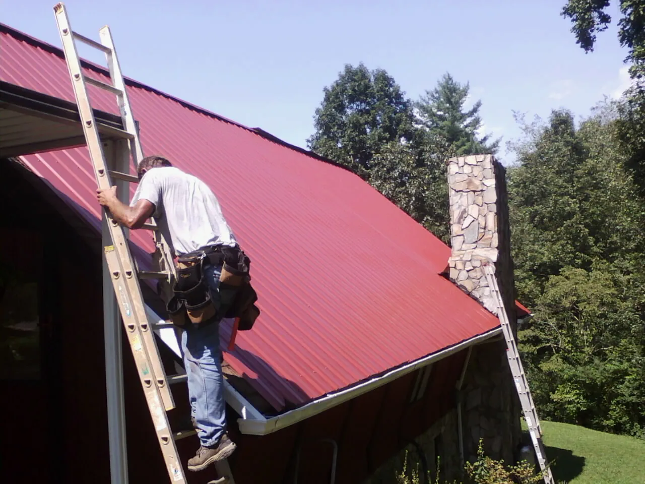 Roofer on a ladder beside a red metal roof and stone chimney — Soffit & Fascia Repair by Roofing Brothers in Thetford Center